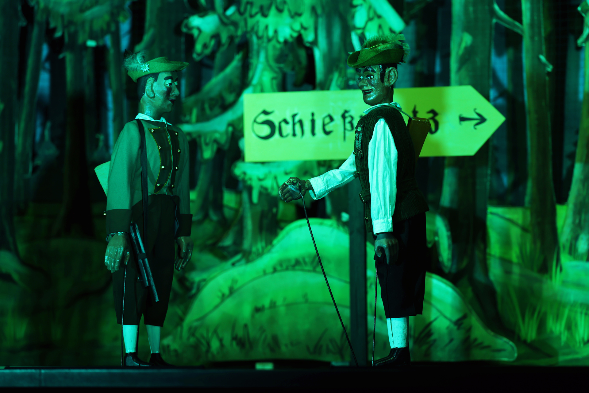 Two stick puppets - the Freischütz with a rifle around his shoulders and the Schängche next to him - stand in front of a "Shooting range" sign. Behind them is a painted backdrop with a fir forest.