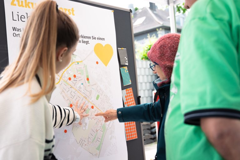 Two women are standing in front of a billboard. One of them is explaining the plans shown on it to the other.