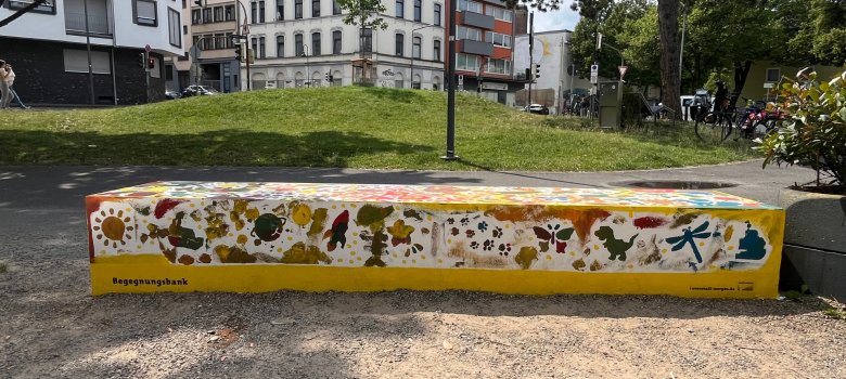 Un banc peint (par exemple soleil, libellule, papillon, dino, pattes d'animaux) en forme de boîte devant une colline d'herbe et des façades de maisons en arrière-plan.