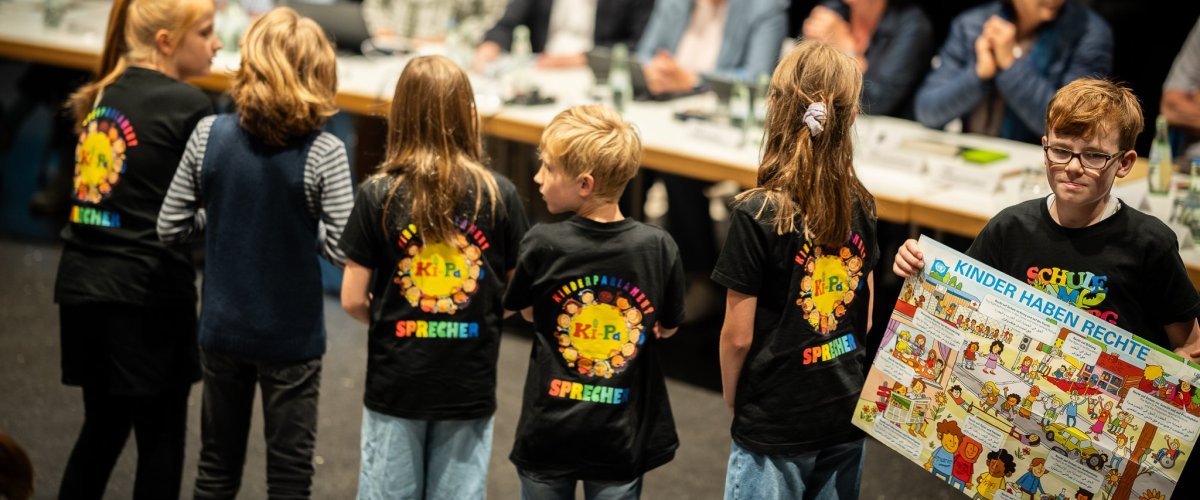 Les enfants portent des t-shirts avec l'inscription "porte-parole du parlement des enfants". Un garçon tient une pancarte sur laquelle est écrit "Les enfants ont des droits". En arrière-plan, on voit les adultes en train de participer à l'événement autour d'une longue table.