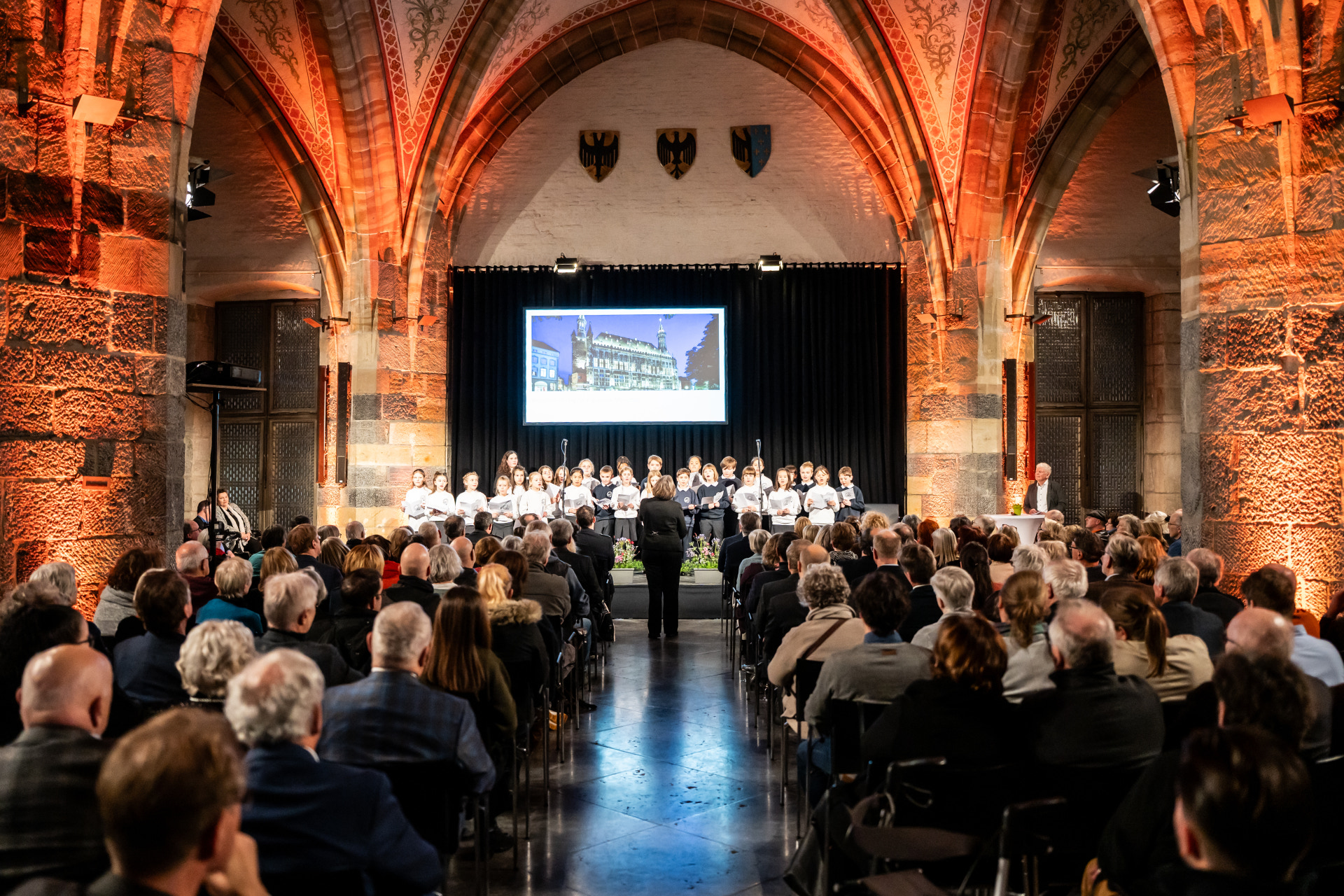 Salle du couronnement pleine à craquer d'invités et, sur scène, un chœur d'enfants et un chef d'orchestre.