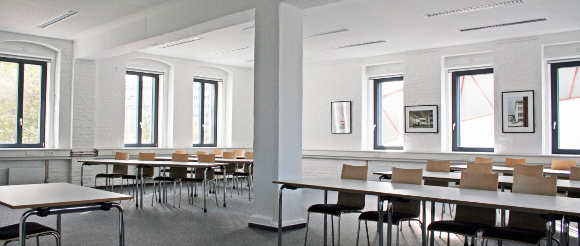 Pièce aux murs blancs et à la moquette grise, ainsi que des tables et des chaises et une colonne blanche au centre.