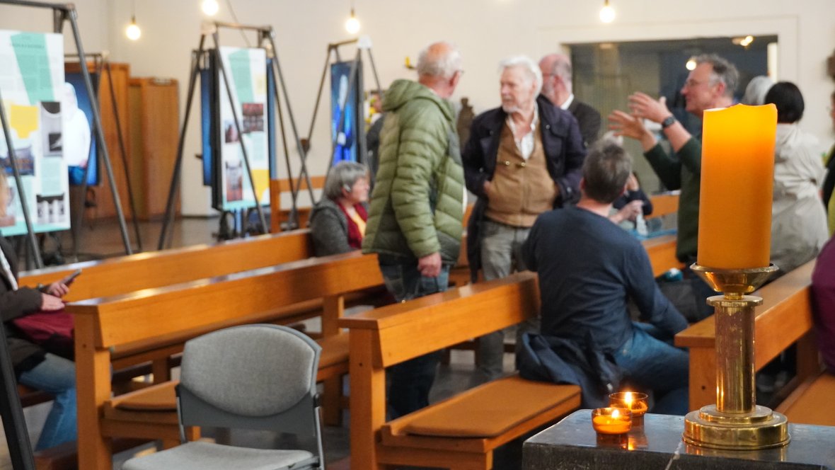 Des visiteurs* discutent devant une exposition sur les églises comme quatrièmes lieux. Au premier plan, trois bougies sont allumées.