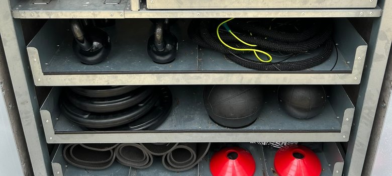 La boîte de sport est ouverte. On voit quatre tiroirs avec différents équipements, comme des ballons, des haltères et des tapis.