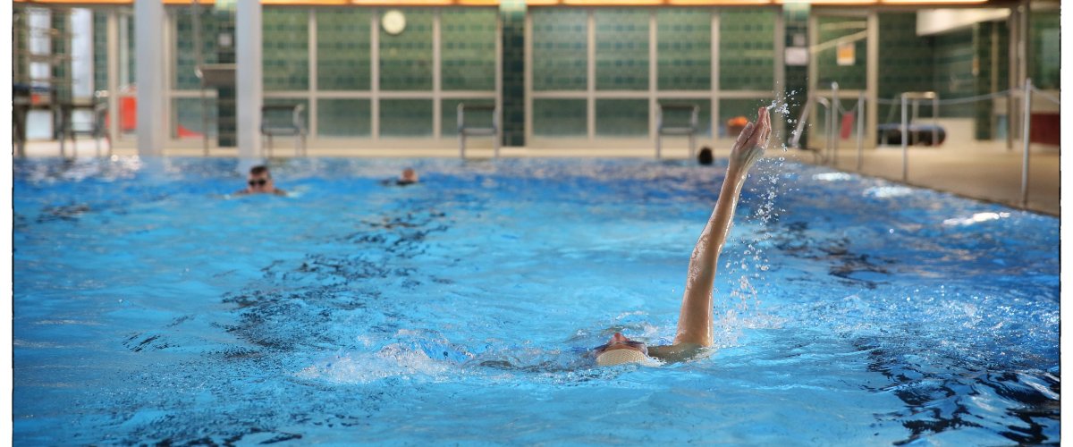 Une nageuse se cramponne sur le dos dans le bassin de la piscine couverte Brand. En arrière-plan, on peut voir la tour de saut.