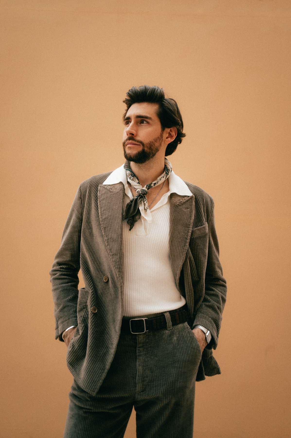Alvaro Soler regarde au loin, les mains dans les poches de son pantalon. Il se tient devant un mur orange.