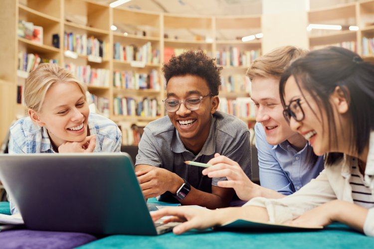 Des étudiants positivement excités, en tenue décontractée, allongés par terre dans la bibliothèque et riant en regardant une vidéo étrange sur leur ordinateur portable.
