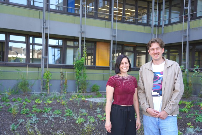 Deux personnes rient face à la caméra, en arrière-plan des plantes vertes devant un immeuble de bureaux.