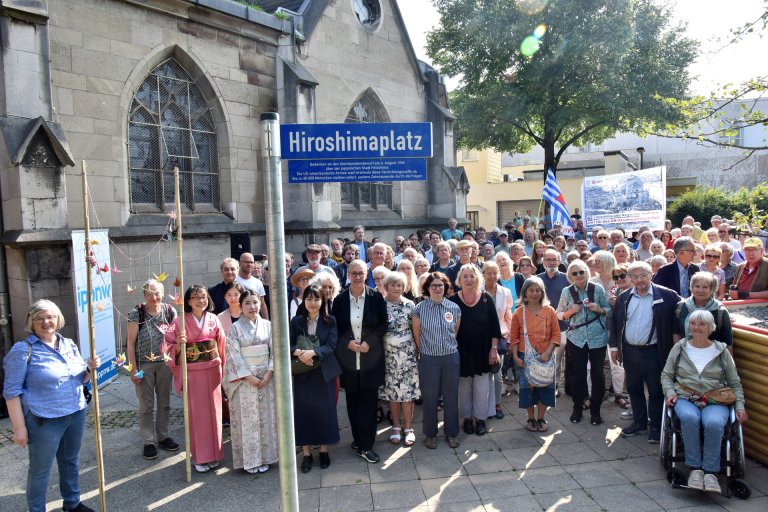 La ville d'Aix-la-Chapelle et l'Alliance contre les armes nucléaires célèbrent l'inauguration de la place d'Hiroshima. Des gens se tiennent derrière un panneau de signalisation.