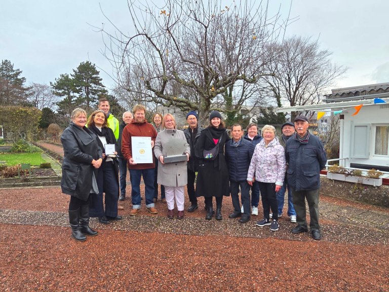 Un groupe de personnes devant une petite maison d'association et des jardins familiaux, ils tiennent un challenge et un certificat.