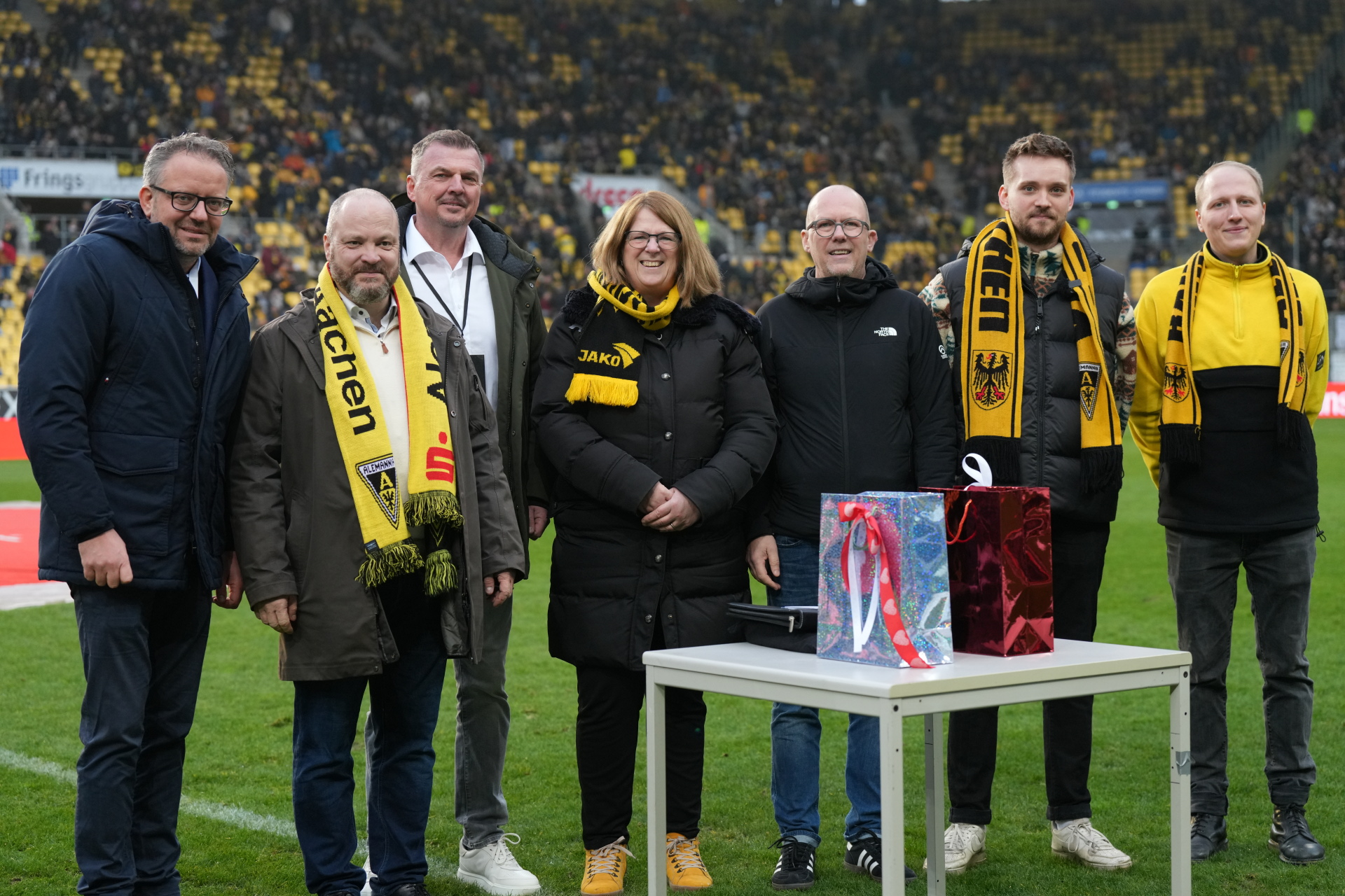 Sept personnes se tiennent autour d'une table dans un stade de football et tirent au sort des billets dans un pot de loterie.