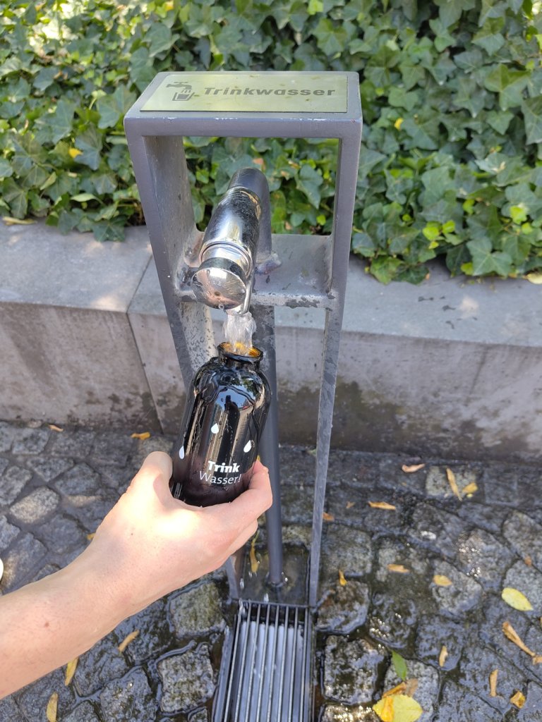 Une bouteille d'eau en métal noir est remplie à une fontaine d'eau potable. Sur la fontaine d'eau potable, il est écrit en noir "eau potable".
