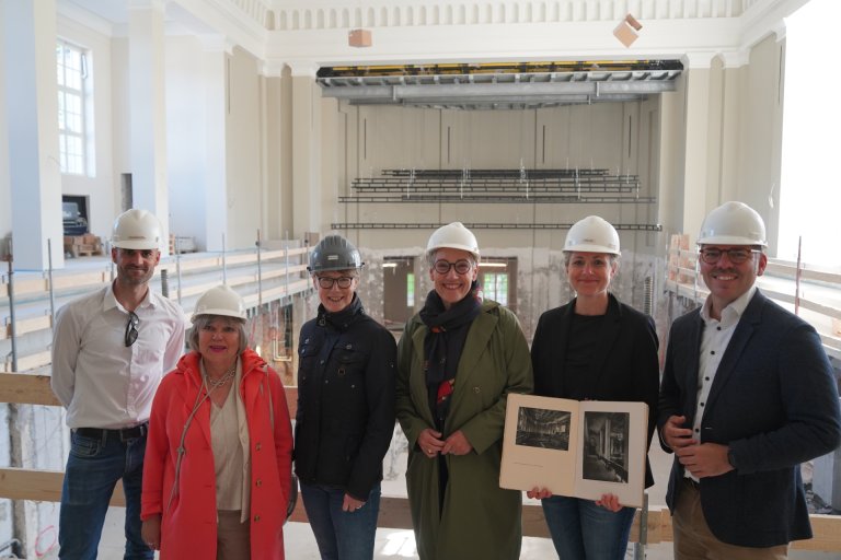 Six personnes, dont le maire, se tiennent sur une galerie dans une grande salle. Celle-ci est encore en chantier.