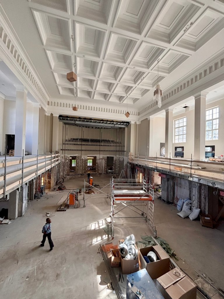 Depuis la galerie, on peut voir la grande salle du nouveau Kurhaus. Il y a du travail sur le chantier. Des artisans se promènent en bas, des câbles pendent du plafond.
