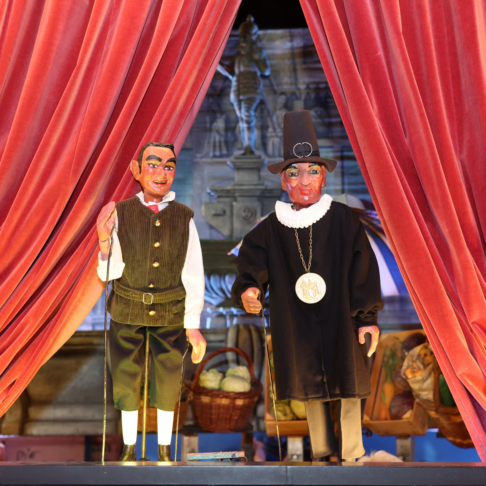 Deux poupées devant une cape rouge sur une scène, côte à côte. La figurine de gauche montre un homme, le Öcher Schängche, celle de droite un homme avec un chapeau et une grande médaille autour du cou.