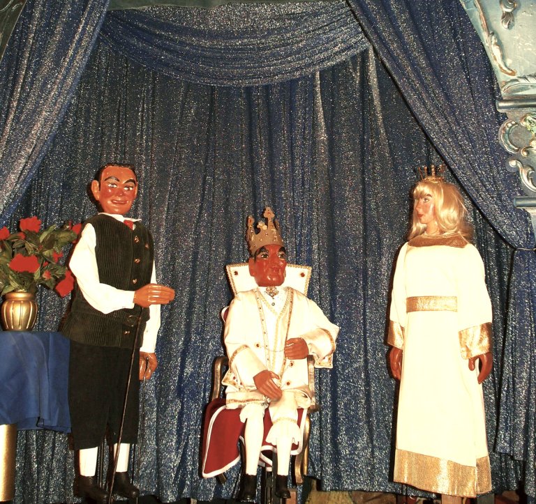 Trois personnages de poupées devant un rideau scintillant : à gauche, un personnage masculin debout, au centre, un roi avec une couronne assis sur le trône, à droite, une femme avec une petite couronne et une robe blanche.