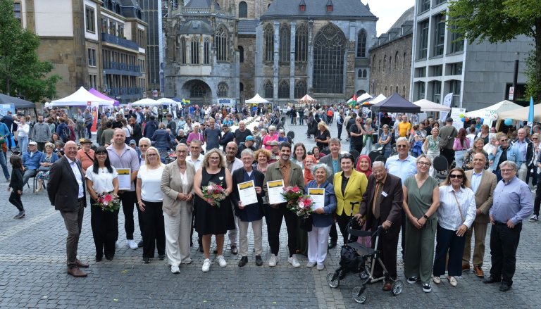 Beaucoup de gens rient dans la caméra. En arrière-plan, une longue table, de nombreuses tentes et stands d'information et la cathédrale d'Aix-la-Chapelle.