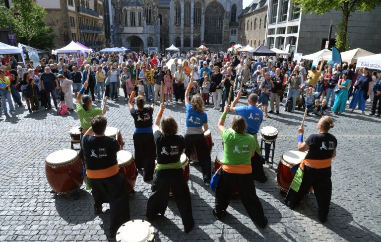 Huit personnes dos à la caméra qui jouent du tambour. Derrière, beaucoup de gens qui regardent. En arrière-plan, une longue table sur une place