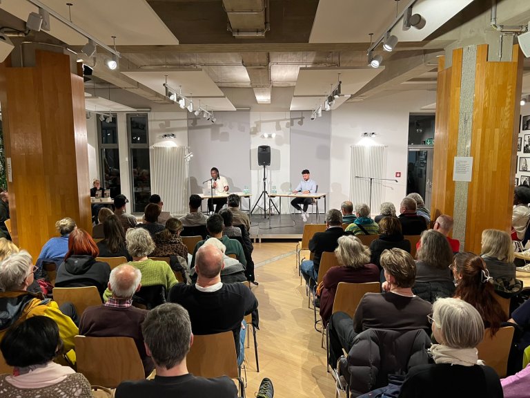 Vue d'une pièce avec beaucoup de gens devant une scène. Sur la scène, deux hommes assis à des tables avec des microphones.