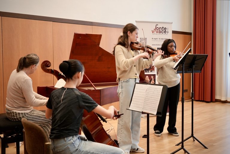 De jeunes musiciens sont debout et assis avec des instruments à cordes devant un piano à queue sur lequel joue une pianiste.