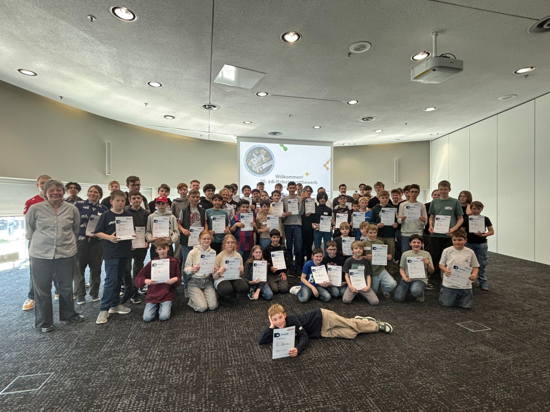 Un grand groupe d'enfants et d'adolescents se tient dans une grande salle. Tous tiennent des diplômes dans les mains.