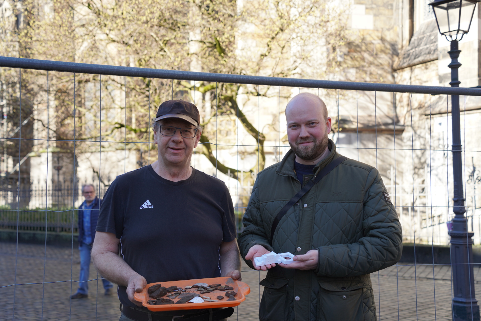 Deux hommes avec de petits objets archéologiques à la main se tiennent devant une clôture de chantier.