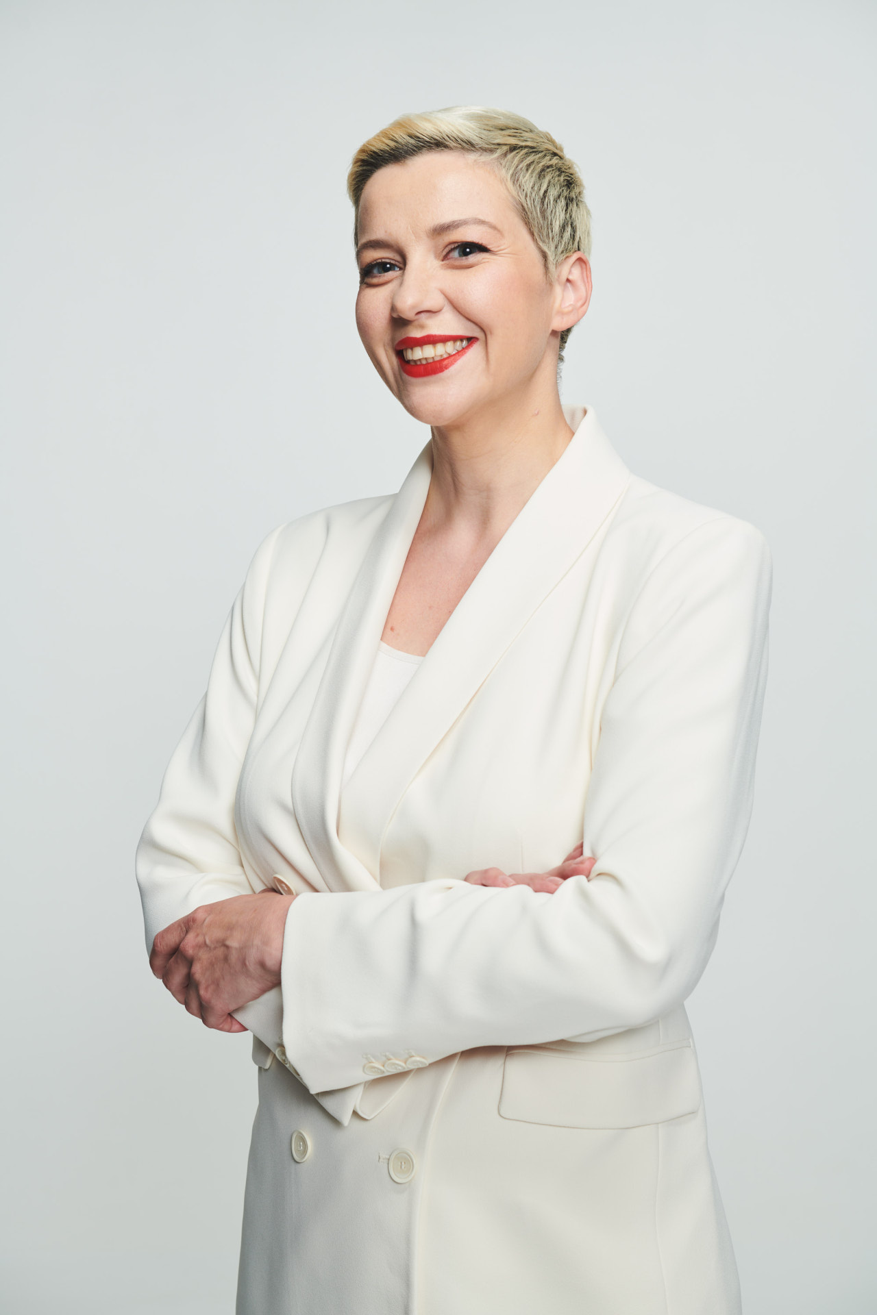 Une femme blonde aux cheveux courts, vêtue d'un costume blanc, sourit à la caméra.