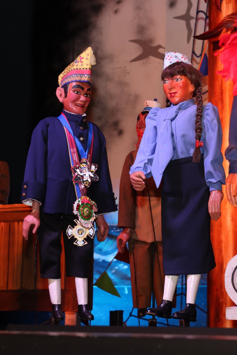 Deux marionnettes à tringle, l'une avec un chapeau de carnaval, l'autre avec des tresses, se tiennent sur un bateau de scène.