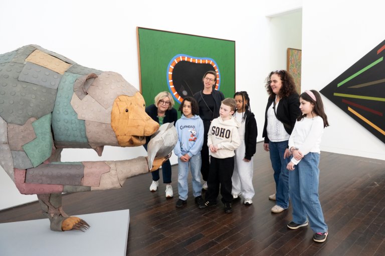Un groupe d'adultes et d'enfants se tient devant une grande sculpture d'ours colorée avec un poisson dans la bouche et la regarde.