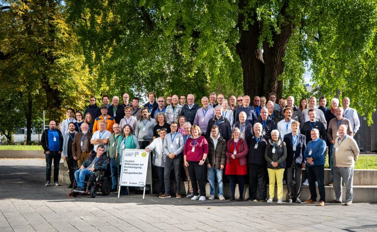 Un grand groupe de personnes est debout sur des marches, autour d'un grand bosquet avec un arbre imposant en arrière-plan.