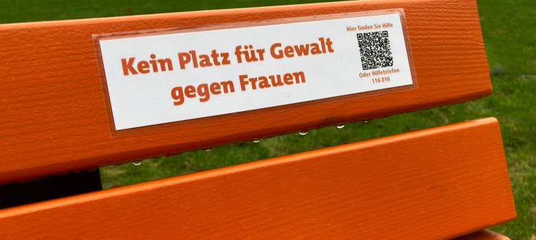 Een oranje bankje met het opschrift "Geen plaats voor geweld tegen vrouwen" en daarnaast een QR-code met de opmerking "Hier kun je hulp vinden of hulptelefoon 116016".