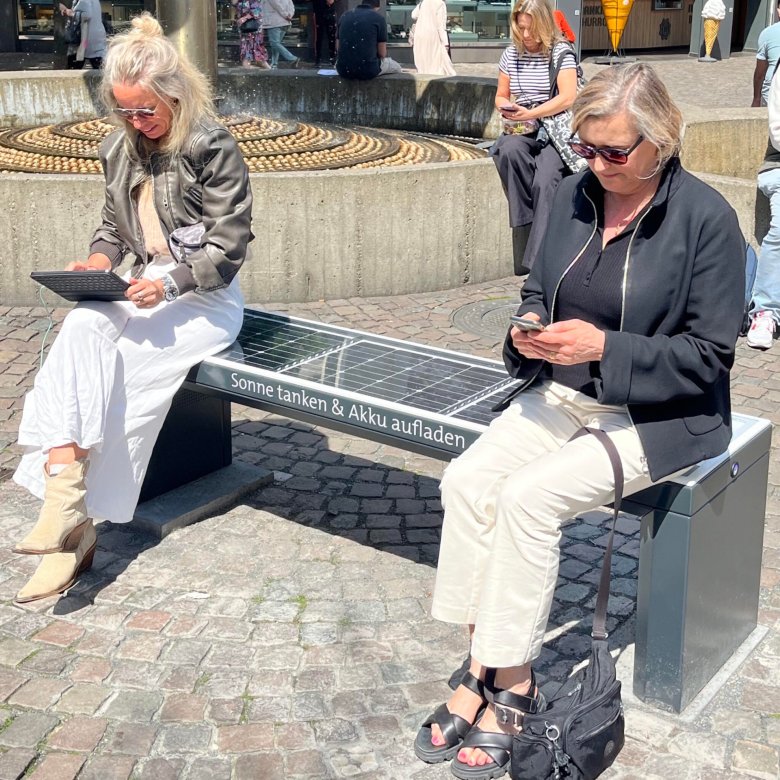 Twee vrouwen zitten op een bankje met een tablet en mobiele telefoon, waarop aan de bovenkant zonnepanelen te zien zijn. Op de voorkant staat geschreven: "Recharge the sun & recharge your battery".