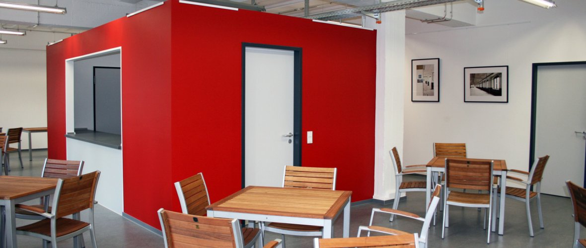 Op de foto zie je bruine tafels en stoelen. Aan de witte muur hangen twee foto's.  In het midden is het keukenblok te zien. De muren zijn rood.