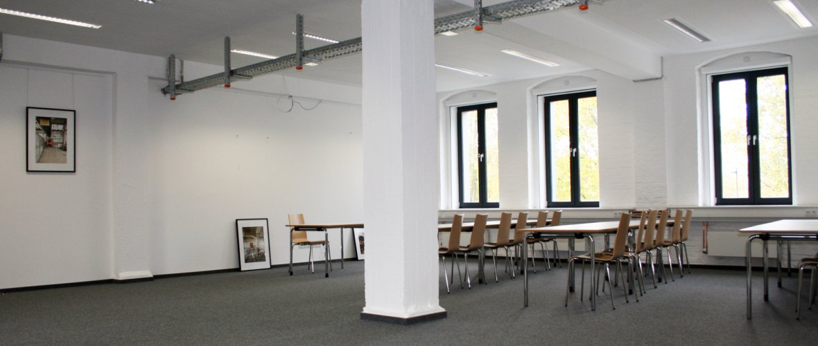 Kamer met witte muren en grijs tapijt, tafels en stoelen en een witte pilaar in het midden.