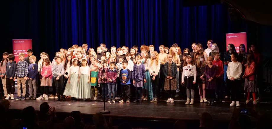 Een grote groep kinderen staat op een groot podium voor een blauw gordijn