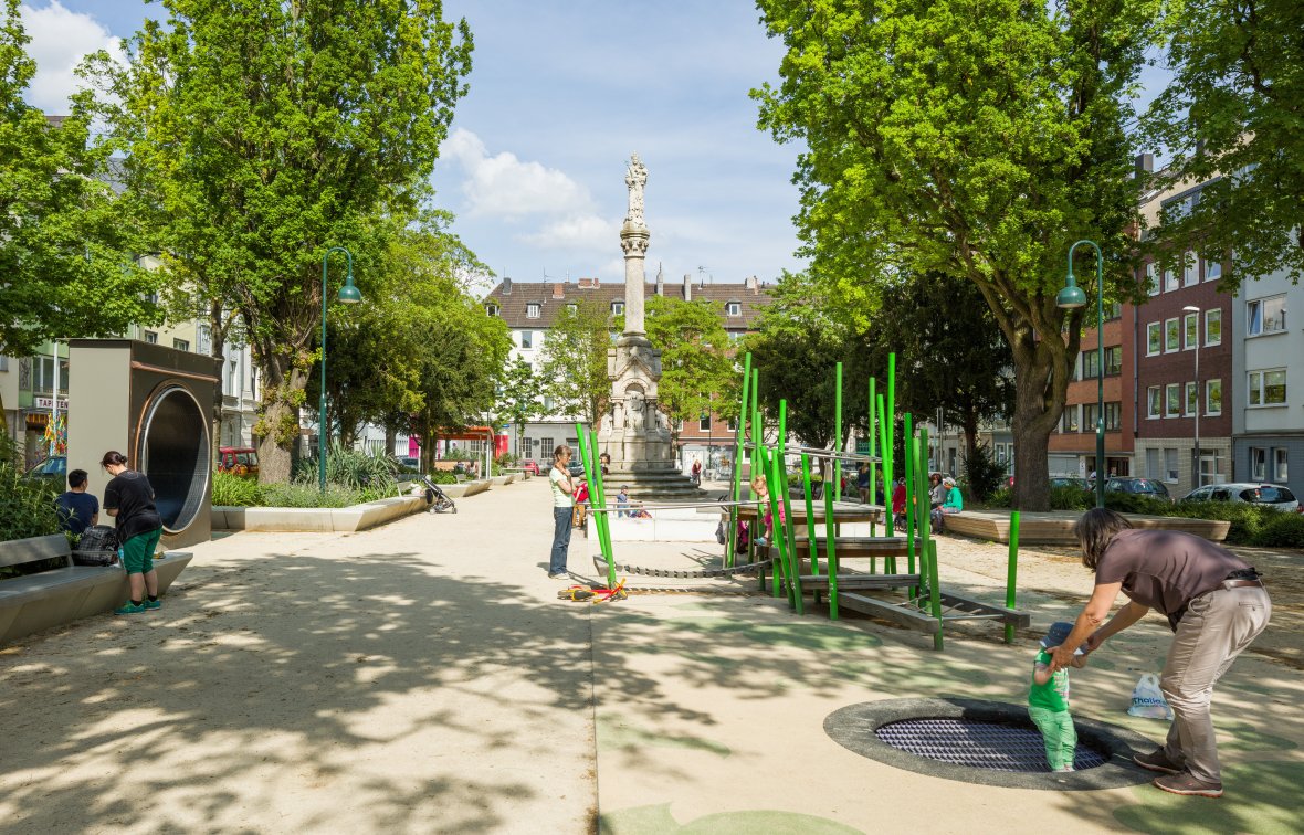 Nieuwe speeltuin met klimtoestellen en veel groen. Kinderen spelen; rechts helpt een vrouw een peuter op een grondtrampoline.