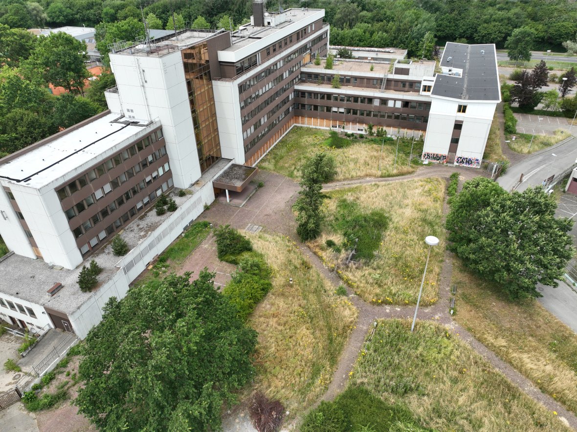 Luchtfoto gemaakt met een drone van het hoofdbureau van politie in Aken. Je ziet het gebouw met meerdere vleugels en een witte en bruine gevel helemaal in beeld. Op de voorgrond ligt een parkachtig groengebied met paden, bomen en grasvelden.