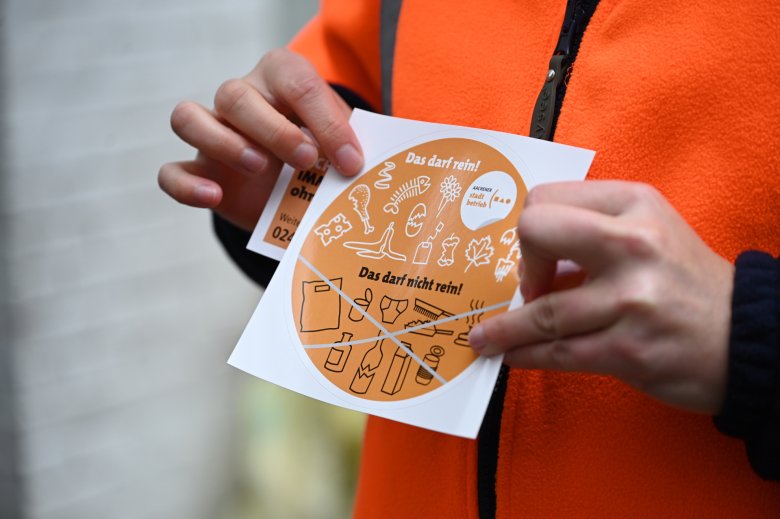 Een oranje sticker met symbolen voor organisch afval wordt vastgehouden door een persoon in oranje kleding. Alleen de handen van de persoon zijn te zien.
