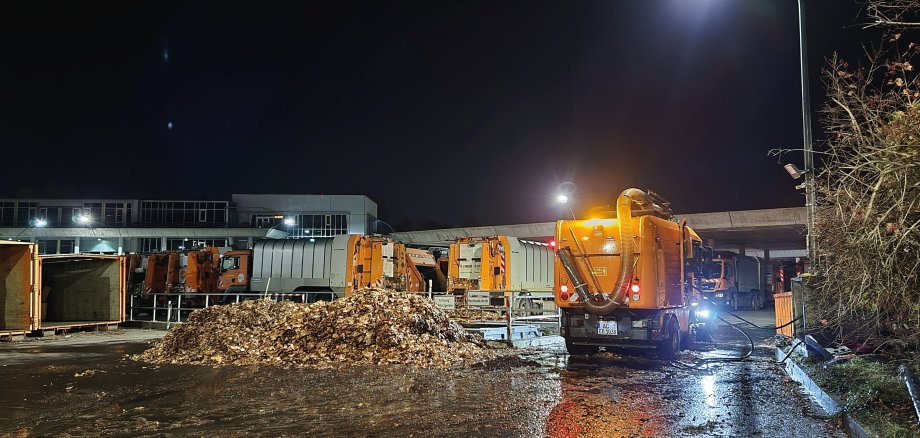 Een straatveger staat in het donker met zijn lichten aan naast een stapel bladeren. Voertuigen voor afvalinzameling op de achtergrond