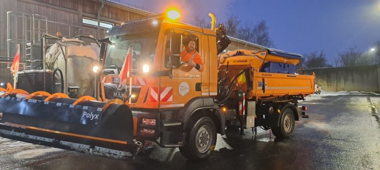 Een winterdienstvoertuig met een sneeuwschuiver en zwaailichten staat in het donker geparkeerd bij een remise. Een man kijkt uit de cabine en steekt zijn duim op.
