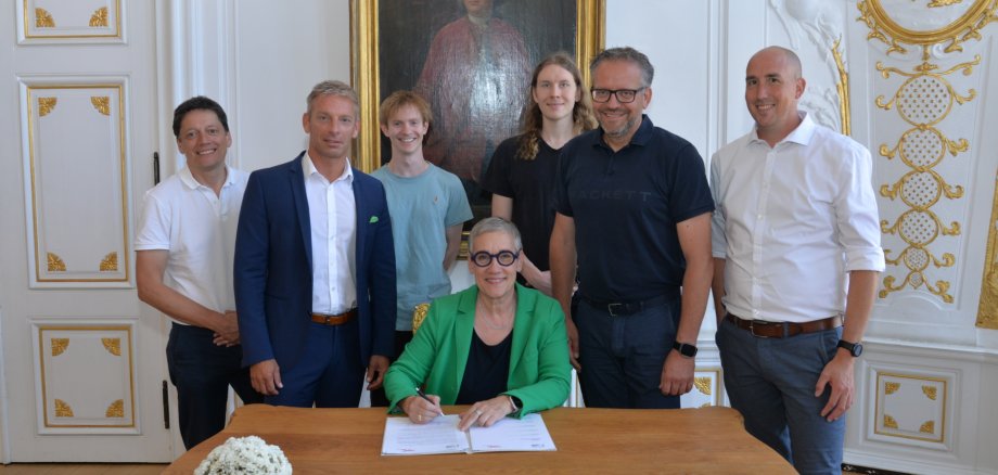 Een groep mensen staat bij een bureau, een vrouw zit op een stoel en ondertekent een document. Op de achtergrond is een witte muur met gouden ornamenten en een historisch schilderij.
