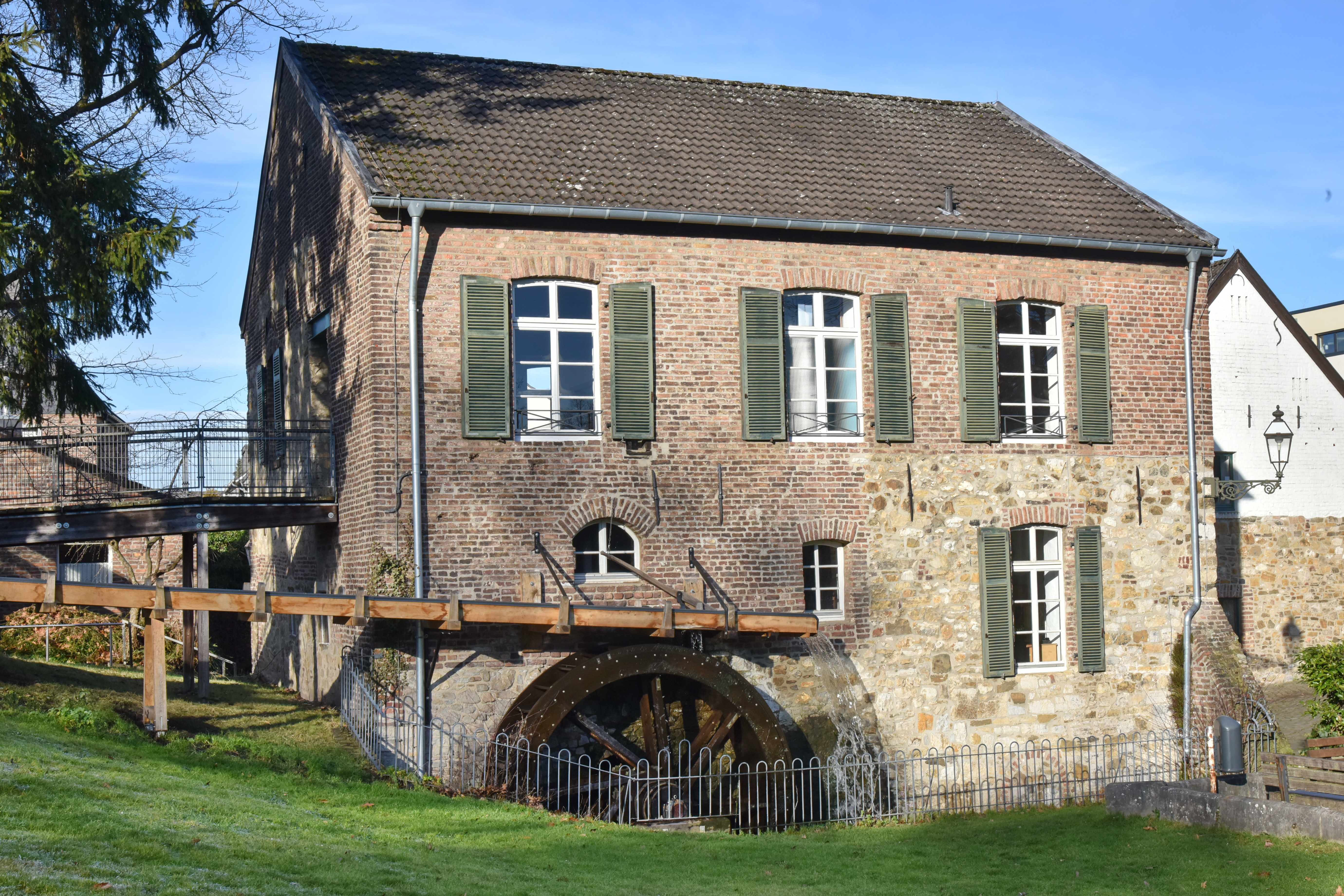De Welsche Mühle onder een blauwe hemel en zonneschijn. Het gebouw is gemaakt van oude bakstenen. De groene luiken staan open.