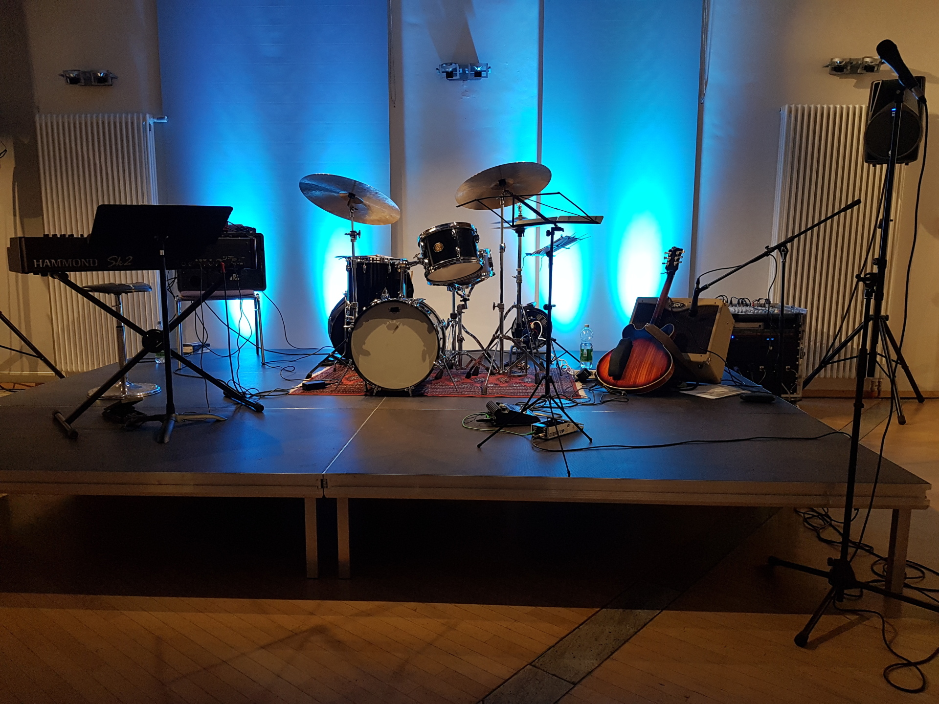 De afbeelding toont een podium met verschillende muziekinstrumenten, zoals een gitaar, een drumstel en een piano. Aan de rechterkant staat een microfoonstandaard met een microfoon. Het podium is verlicht in de kleur blauw.