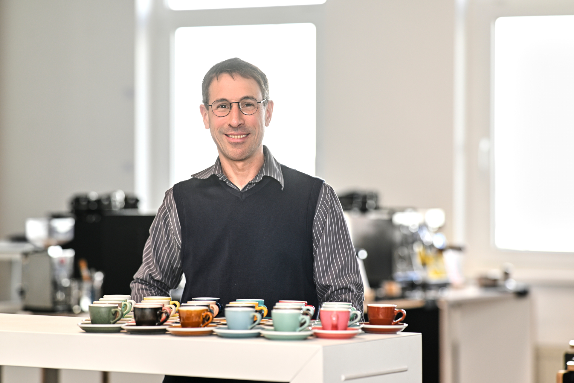 Een man staat in een kamer voor een tafel waarop kleurrijke espressokopjes staan.