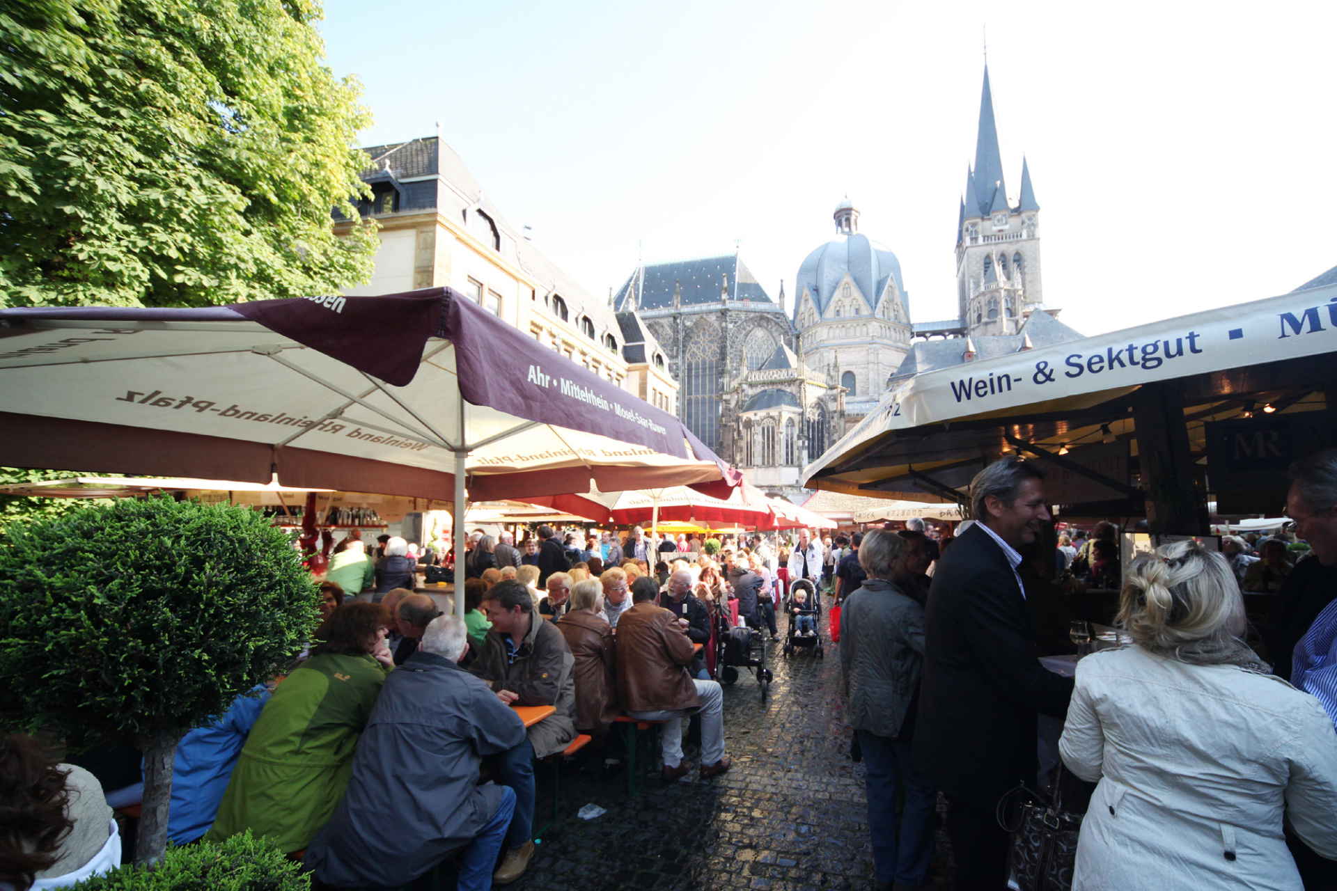 Mensen zitten onder parasols op bankjes bij Katschhof en drinken wijn. Het is zomer en de zon schijnt.