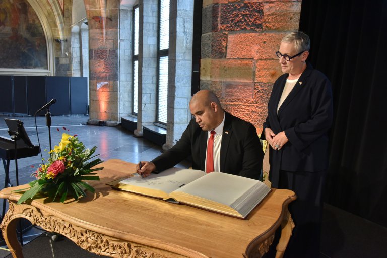 Eddie Andrews, de loco-burgemeester van Kaapstad, schrijft in het Gouden Boek van Aken, zittend aan een bureau, terwijl de burgemeester van Aken, Sibylle Keupen, rechts van hem staat.