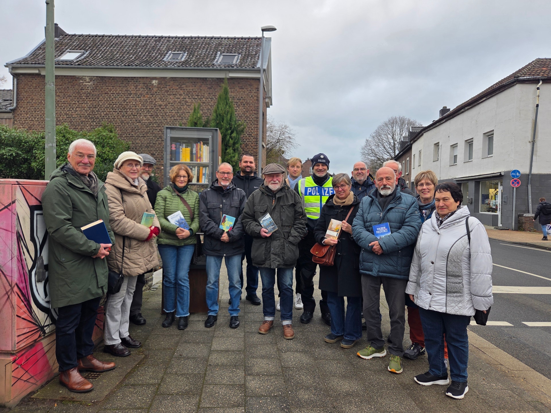 Een groep mensen met boeken in hun handen staat naast een boekenkast op een stoep.