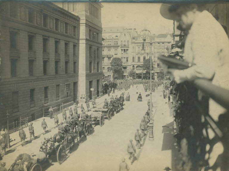 Historische foto: Paardenkoetsen, soldaten en andere mensen passeren de noordkant van de stadsschouwburg. Veel mensen op straat en in de ramen van de aangrenzende gebouwen