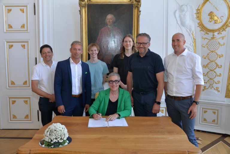 Een groep mensen staat voor een houten bureau. Een vrouw zit op een stoel bij het bureau en ondertekent een vel papier. Op de achtergrond is een witte muur met gouden ornamenten en een historisch schilderij.