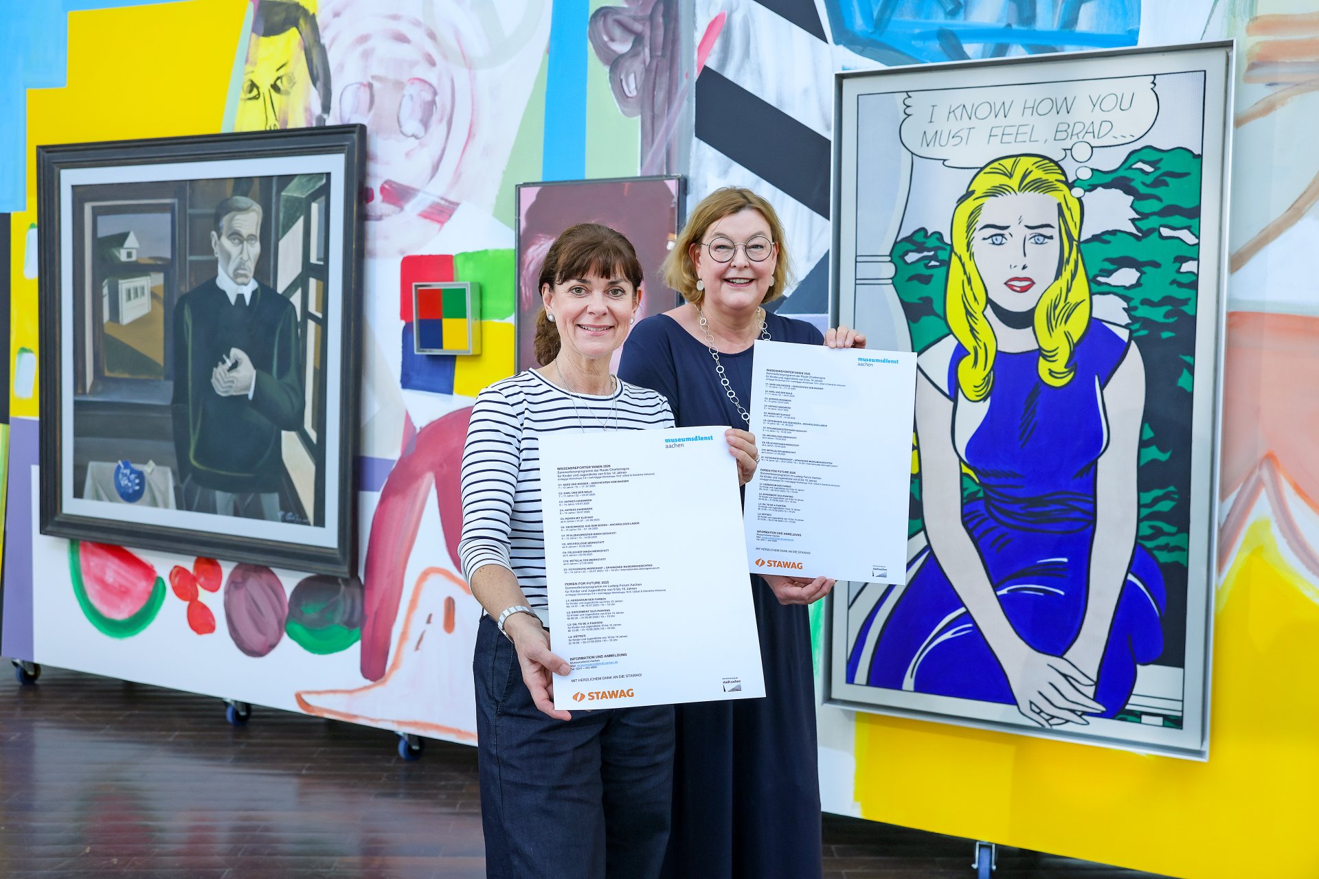 Twee vrouwen staan voor een kleurrijke kunstmuur en houden posters omhoog.